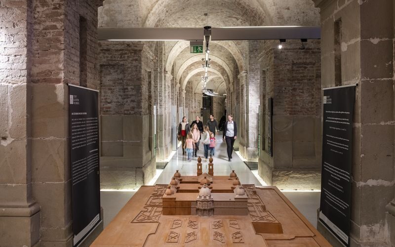 Eine Gruppe von Personen geht durch einen gewölbten Steinflur, in dessen Vordergrund Ausstellungstafeln und ein hölzernes Architekturmodell zu sehen sind.