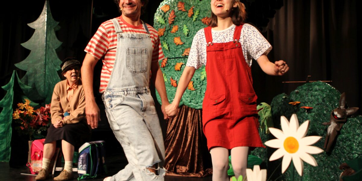 Zwei Personen in Overalls halten sich an den Händen und lächeln, während sie über eine mit Bäumen und Blumen geschmückte Bühne gehen, während eine weitere Person im Hintergrund sitzt.