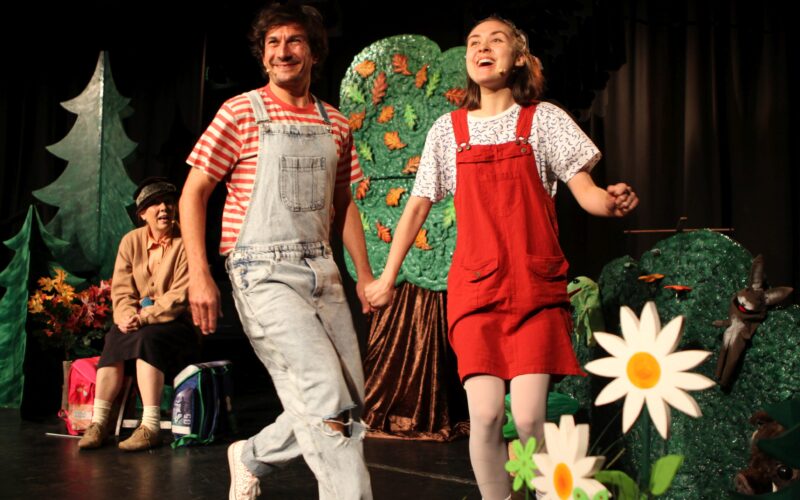 Zwei Personen in Overalls halten sich an den Händen und lächeln, während sie über eine mit Bäumen und Blumen geschmückte Bühne gehen, während eine weitere Person im Hintergrund sitzt.