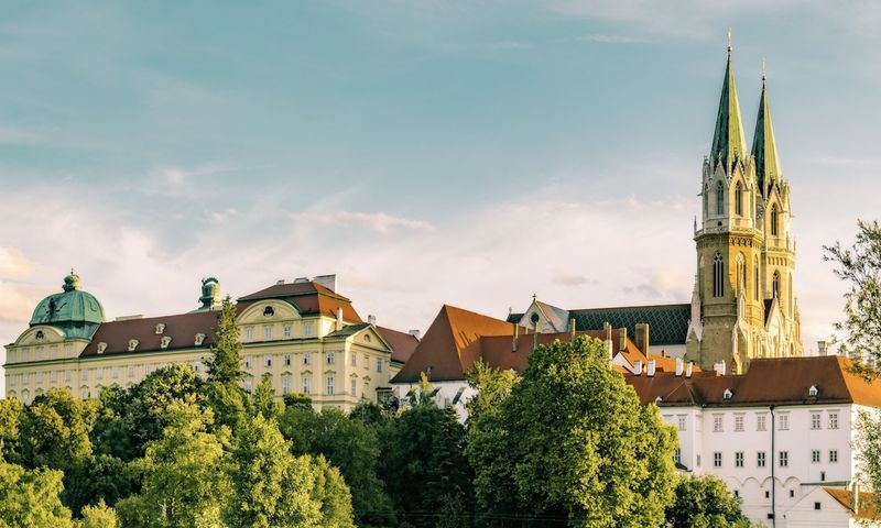 Ein großer historischer Gebäudekomplex mit einer hohen Kirche mit zwei spitzen Türmen, umgeben von Bäumen unter einem klaren Himmel.