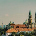 Große Kirche mit Zwillingstürmen und grünen Kuppeln, umgeben von Gebäuden mit roten Dächern, mit einer modernen Stadtsilhouette im Hintergrund.