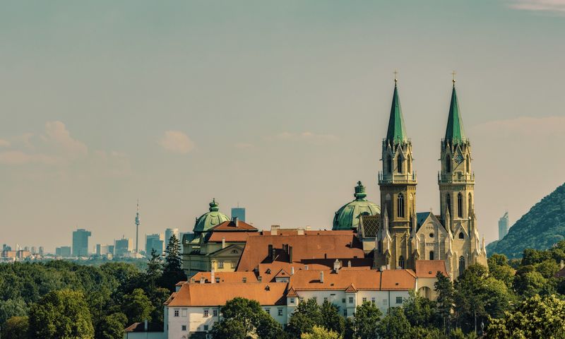Große Kirche mit Zwillingstürmen und grünen Kuppeln, umgeben von Gebäuden mit roten Dächern, mit einer modernen Stadtsilhouette im Hintergrund.
