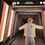 Ein Kind steht mit ausgestreckten Armen in einem Tunnel an der Grenze zwischen Österreich und Deutschland, unter einem Schild mit der Aufschrift "Internationale Grenze" und umgeben von beleuchteten österreichischen Fahnen.