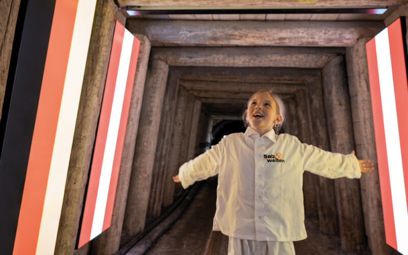 Ein Kind steht mit ausgestreckten Armen in einem Tunnel an der Grenze zwischen Österreich und Deutschland, unter einem Schild mit der Aufschrift "Internationale Grenze" und umgeben von beleuchteten österreichischen Fahnen.