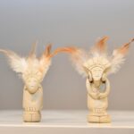 Zwei kleine elfenbeinfarbene Figuren mit detaillierten Schnitzereien und Federkopfschmuck sind auf einer weißen Fläche vor einem hellen Hintergrund ausgestellt.