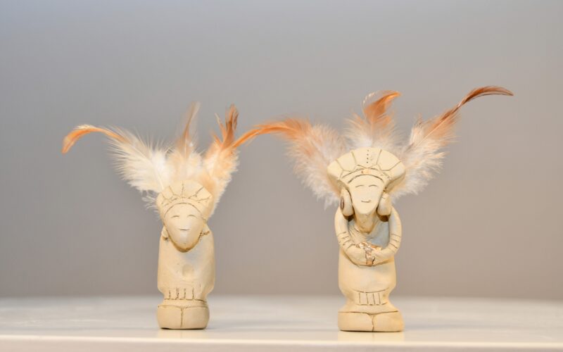 Zwei kleine elfenbeinfarbene Figuren mit detaillierten Schnitzereien und Federkopfschmuck sind auf einer weißen Fläche vor einem hellen Hintergrund ausgestellt.