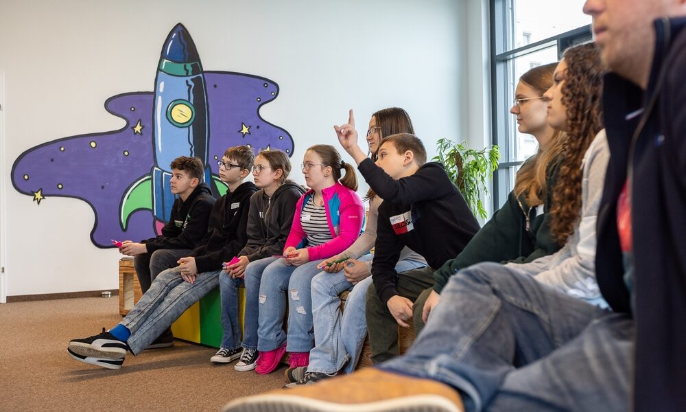 Eine Gruppe junger Menschen sitzt auf Bänken in einem Innenraum, einige heben die Hände, vor einer Wandmalerei, die eine Cartoon-Rakete und Sterne zeigt.