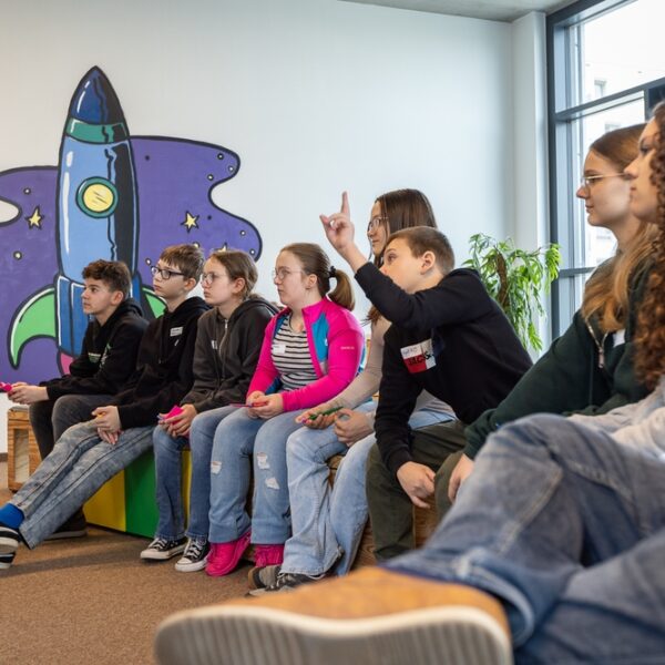 Eine Gruppe junger Menschen sitzt auf Bänken in einem Innenraum, einige heben die Hände, vor einer Wandmalerei, die eine Cartoon-Rakete und Sterne zeigt.