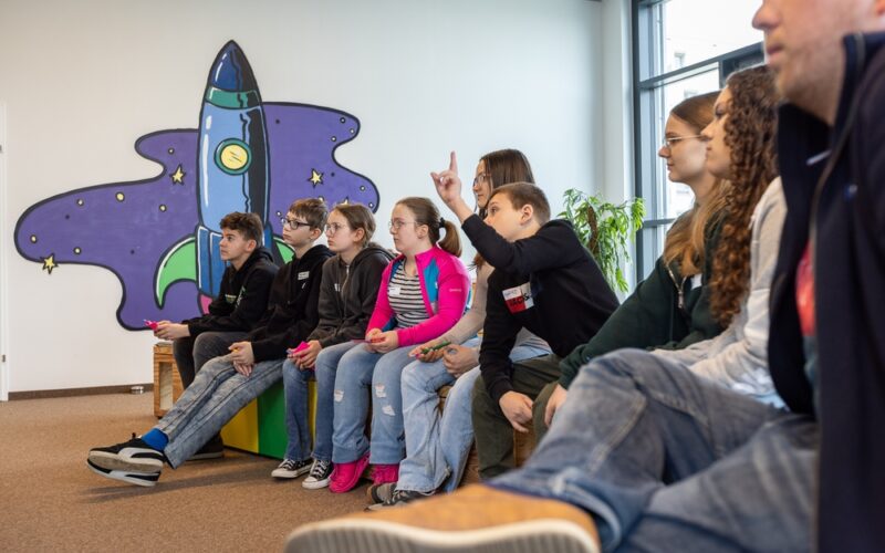 Eine Gruppe junger Menschen sitzt auf Bänken in einem Innenraum, einige heben die Hände, vor einer Wandmalerei, die eine Cartoon-Rakete und Sterne zeigt.