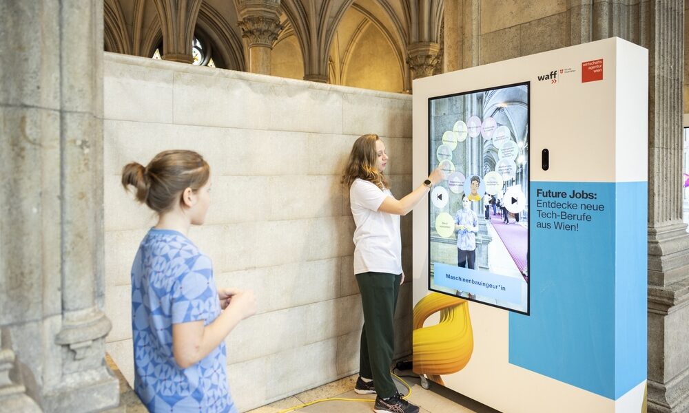 Zwei Personen interagieren mit einem großen Touchscreen-Display, das in einem historischen Gebäude mit gewölbter Steinarchitektur aufgestellt ist und über künftige Arbeitsplätze in der Technologiebranche in Wien informiert.