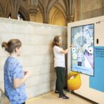 Zwei Personen interagieren mit einem großen Touchscreen-Display, das in einem historischen Gebäude mit gewölbter Steinarchitektur aufgestellt ist und über künftige Arbeitsplätze in der Technologiebranche in Wien informiert.