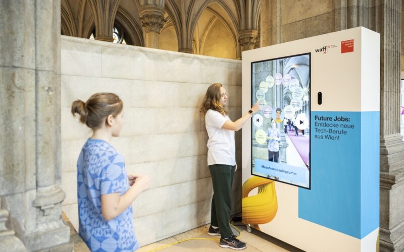Zwei Personen interagieren mit einem großen Touchscreen-Display, das in einem historischen Gebäude mit gewölbter Steinarchitektur aufgestellt ist und über künftige Arbeitsplätze in der Technologiebranche in Wien informiert.