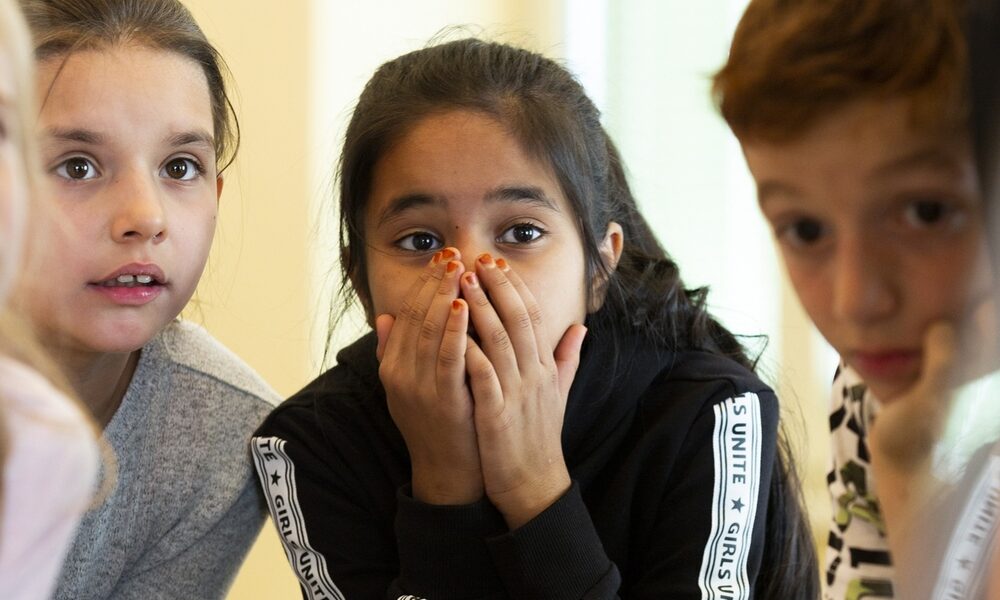 Eine Gruppe von Kindern versammelt sich um einen Tisch. Ein Mädchen in der Mitte hält sich die Hände vor den Mund und wirkt überrascht oder erstaunt.