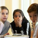 Eine Gruppe von Kindern versammelt sich um einen Tisch. Ein Mädchen in der Mitte hält sich die Hände vor den Mund und wirkt überrascht oder erstaunt.