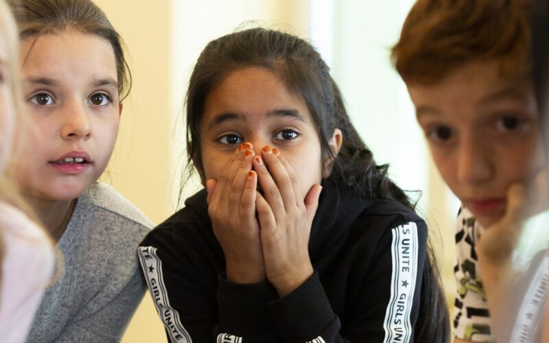 Eine Gruppe von Kindern versammelt sich um einen Tisch. Ein Mädchen in der Mitte hält sich die Hände vor den Mund und wirkt überrascht oder erstaunt.