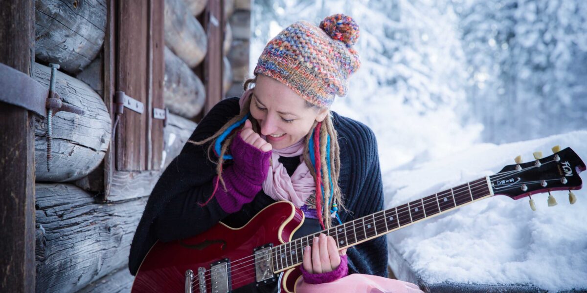 Eine Person, die eine bunte Strickmütze und Handschuhe trägt, hält eine rote E-Gitarre, während sie vor einer rustikalen Holzhütte in einer verschneiten Landschaft sitzt.