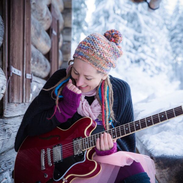 Eine Person, die eine bunte Strickmütze und Handschuhe trägt, hält eine rote E-Gitarre, während sie vor einer rustikalen Holzhütte in einer verschneiten Landschaft sitzt.
