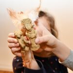 Ein Kind hält zwei kleine, handgefertigte, mit Federn geschmückte Tonfiguren hoch, deren Gesicht im Hintergrund teilweise verdeckt ist.