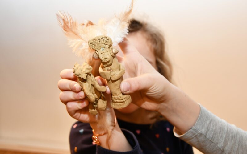 Ein Kind hält zwei kleine, handgefertigte, mit Federn geschmückte Tonfiguren hoch, deren Gesicht im Hintergrund teilweise verdeckt ist.