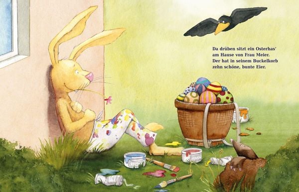 Ein Osterhase sitzt an einer Hauswand und bemalt bunte Eier neben einem Korb; ein schwarzer Vogel schaut von oben zu. Farbdosen und Pinsel liegen auf dem Rasen.