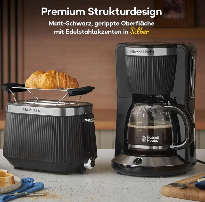 Ein schwarzer Russell Hobbs-Toaster mit einem Croissant und eine passende Kaffeemaschine mit Glaskanne stehen auf einem Küchentresen, daneben liegen Brot und Butter.