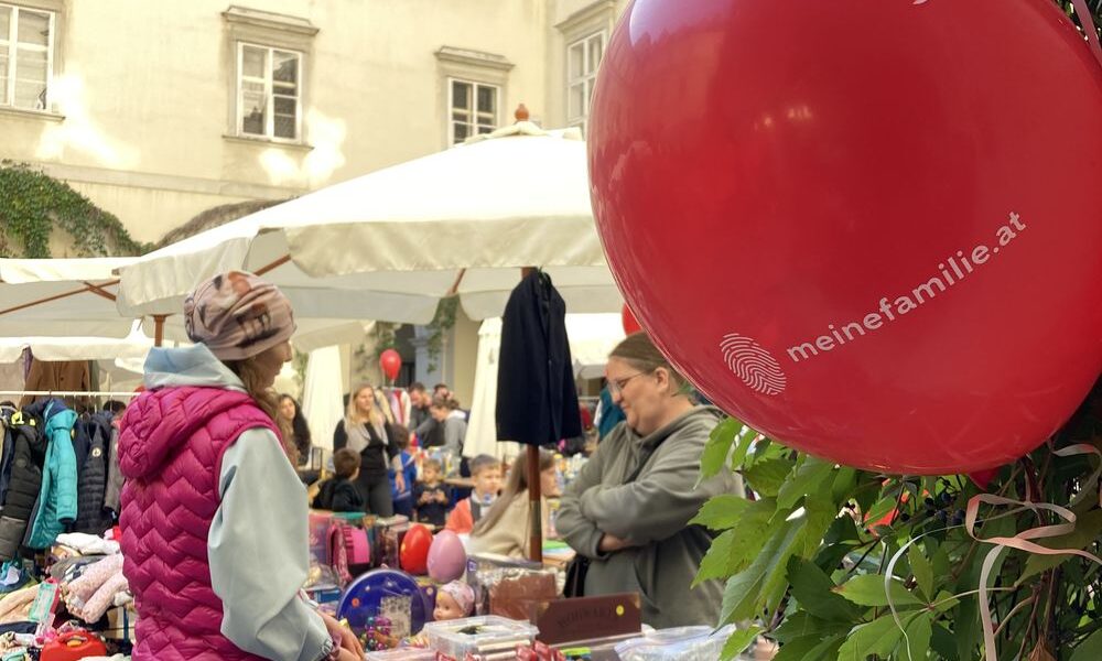 Menschen stöbern auf einem Markt im Freien, auf dem Tische mit Waren zum Verkauf angeboten werden. Ein roter Luftballon mit dem Aufdruck "meinefamilie.at" ist im Vordergrund an Grünzeug gebunden.