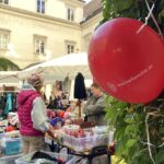 Menschen stöbern auf einem Markt im Freien, auf dem Tische mit Waren zum Verkauf angeboten werden. Ein roter Luftballon mit dem Aufdruck "meinefamilie.at" ist im Vordergrund an Grünzeug gebunden.