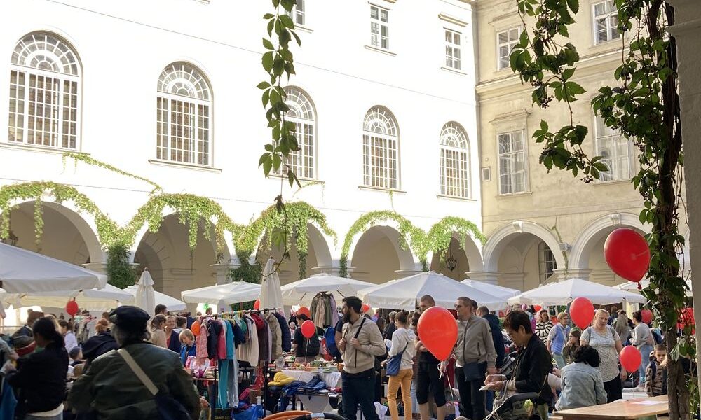 Menschen stöbern an Marktständen in einem Innenhof mit bogenförmigen Gängen. Mehrere Erwachsene schieben Kinderwagen und einige Kinder halten rote Luftballons. Der Himmel ist klar und blau.