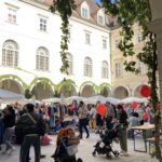 Menschen stöbern an Marktständen in einem Innenhof mit bogenförmigen Gängen. Mehrere Erwachsene schieben Kinderwagen und einige Kinder halten rote Luftballons. Der Himmel ist klar und blau.