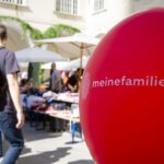 Ein roter Ballon mit dem Text "meinefamilie.at" auf einem Markt im Freien; im Hintergrund sind Menschen und Tische mit Waren zu sehen.