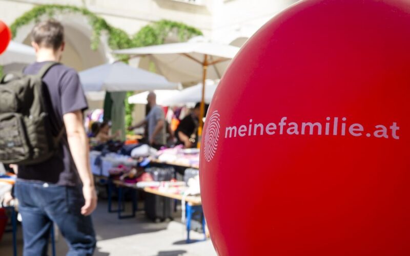 Ein roter Ballon mit dem Text "meinefamilie.at" auf einem Markt im Freien; im Hintergrund sind Menschen und Tische mit Waren zu sehen.