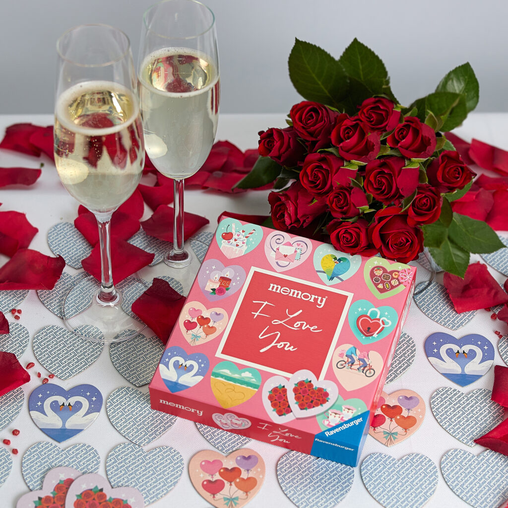 Eine Schachtel mit "I Love You"-Gedächtniskarten, zwei Gläser Sekt und ein Strauß roter Rosen auf einem Tisch mit herzförmigen Dekorationen und Rosenblättern.