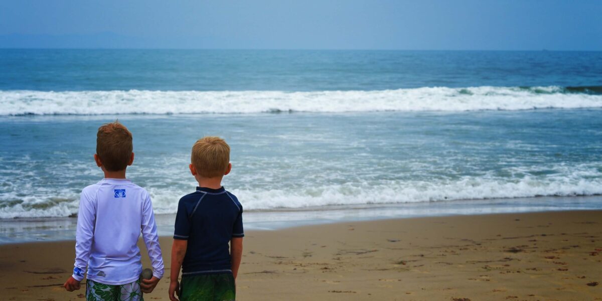 Zwei Jungen in Badekleidung stehen nebeneinander an einem Sandstrand und blicken auf die Wellen des Ozeans unter einem bewölkten Himmel.