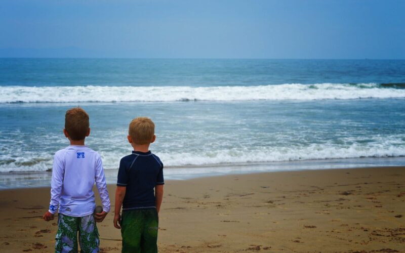 Zwei Jungen in Badekleidung stehen nebeneinander an einem Sandstrand und blicken auf die Wellen des Ozeans unter einem bewölkten Himmel.