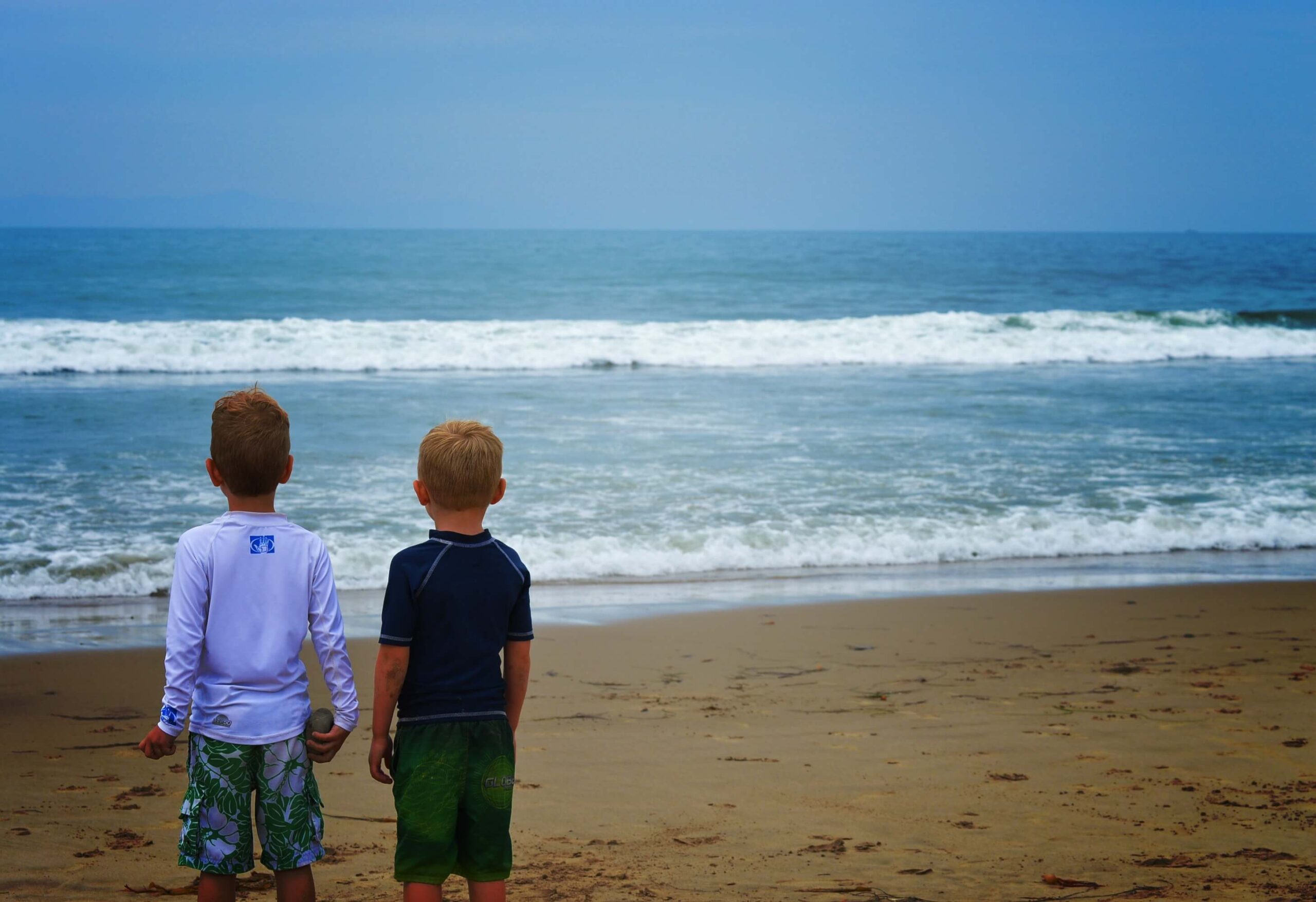 Zwei Jungen in Badekleidung stehen nebeneinander an einem Sandstrand und blicken auf die Wellen des Ozeans unter einem bewölkten Himmel.