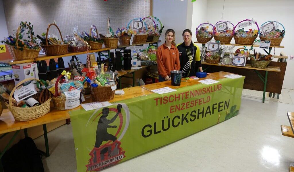 Zwei Personen stehen bei einer Veranstaltung des Tischtennisklubs Enzesfeld hinter einem Tisch mit Tombola-Körben, auf denen ein Banner mit der Aufschrift GLÜCKSHAFEN" und verschiedene Preise zu sehen sind.