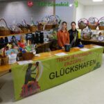 Zwei Personen stehen bei einer Veranstaltung des Tischtennisklubs Enzesfeld hinter einem Tisch mit Tombola-Körben, auf denen ein Banner mit der Aufschrift GLÜCKSHAFEN" und verschiedene Preise zu sehen sind.