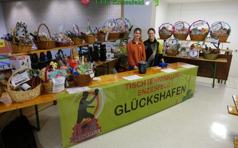 Zwei Personen stehen bei einer Veranstaltung des Tischtennisklubs Enzesfeld hinter einem Tisch mit Tombola-Körben, auf denen ein Banner mit der Aufschrift GLÜCKSHAFEN" und verschiedene Preise zu sehen sind.