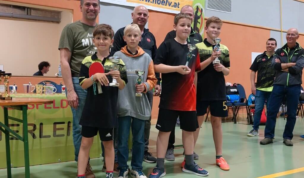 Vier Jungen halten Trophäen und einen Tischtennisschläger in der Hand und posieren mit drei Erwachsenen in einer Turnhalle während einer Preisverleihung.