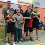 Vier Jungen halten Trophäen und einen Tischtennisschläger in der Hand und posieren mit drei Erwachsenen in einer Turnhalle während einer Preisverleihung.