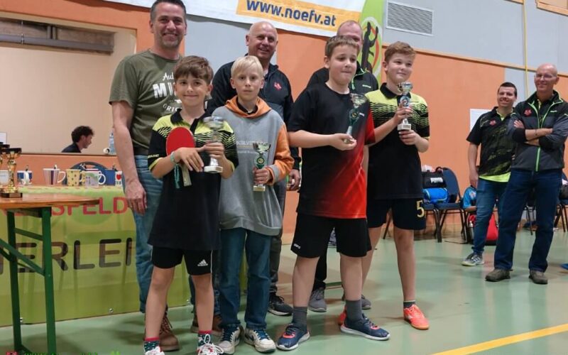 Vier Jungen halten Trophäen und einen Tischtennisschläger in der Hand und posieren mit drei Erwachsenen in einer Turnhalle während einer Preisverleihung.