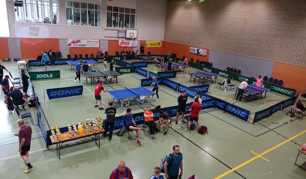 Tischtennisturnier in einer Sporthalle mit mehreren laufenden Spielen, Zuschauern in der Nähe und Trophäen auf einem Tisch im Vordergrund.