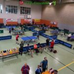 Tischtennisturnier in einer Sporthalle mit mehreren laufenden Spielen, Zuschauern in der Nähe und Trophäen auf einem Tisch im Vordergrund.