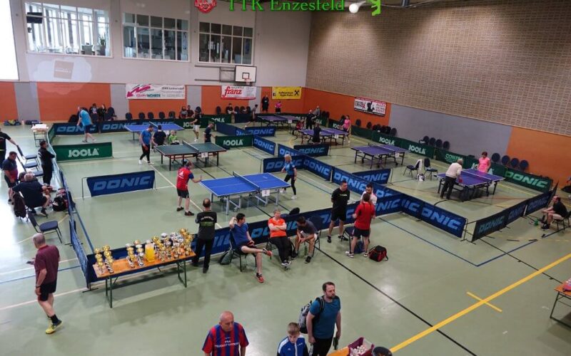 Tischtennisturnier in einer Sporthalle mit mehreren laufenden Spielen, Zuschauern in der Nähe und Trophäen auf einem Tisch im Vordergrund.