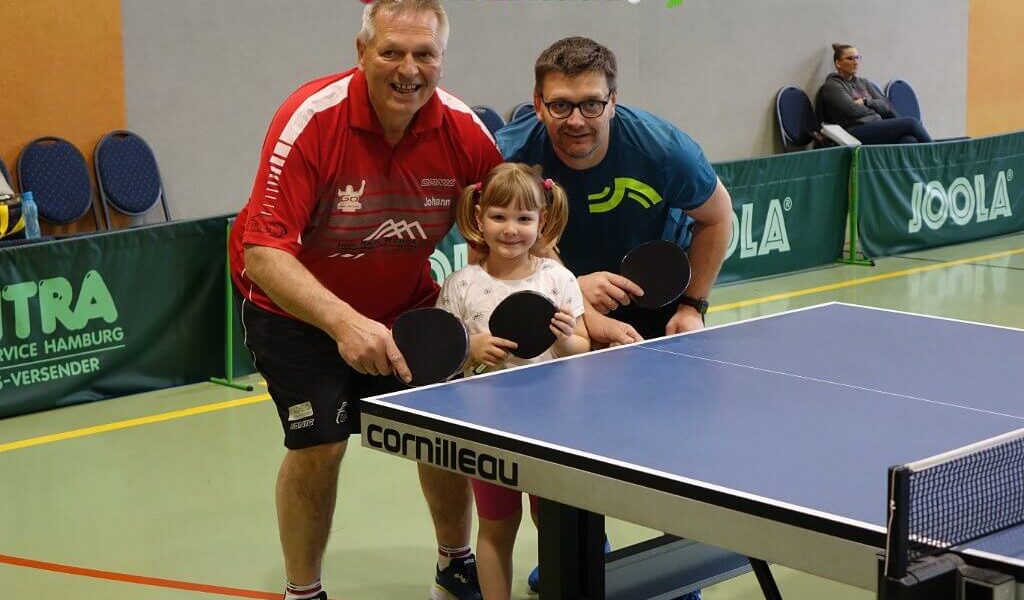 Drei Personen, zwei Erwachsene und ein Kind, posieren mit Tischtennisschlägern neben einer Indoor-Tischtennisplatte in einer Sporthalle.