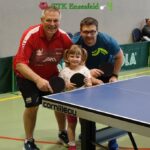 Drei Personen, zwei Erwachsene und ein Kind, posieren mit Tischtennisschlägern neben einer Indoor-Tischtennisplatte in einer Sporthalle.