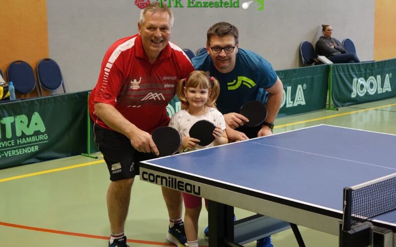 Drei Personen, zwei Erwachsene und ein Kind, posieren mit Tischtennisschlägern neben einer Indoor-Tischtennisplatte in einer Sporthalle.
