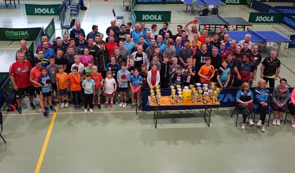 Eine große Gruppe von Menschen, darunter Kinder und Erwachsene, posieren gemeinsam in einer Turnhalle hinter einem Tisch mit Trophäen, umgeben von Tischtennisplatten.