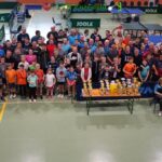 Eine große Gruppe von Menschen, darunter Kinder und Erwachsene, posieren gemeinsam in einer Turnhalle hinter einem Tisch mit Trophäen, umgeben von Tischtennisplatten.