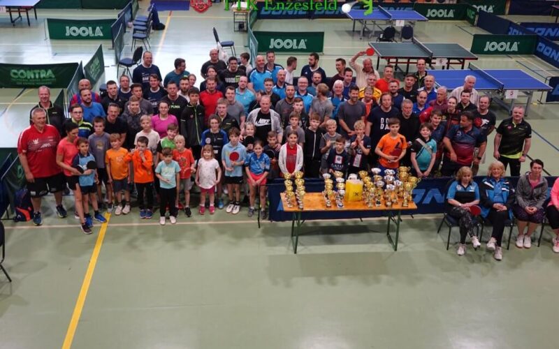 Eine große Gruppe von Menschen, darunter Kinder und Erwachsene, posieren gemeinsam in einer Turnhalle hinter einem Tisch mit Trophäen, umgeben von Tischtennisplatten.